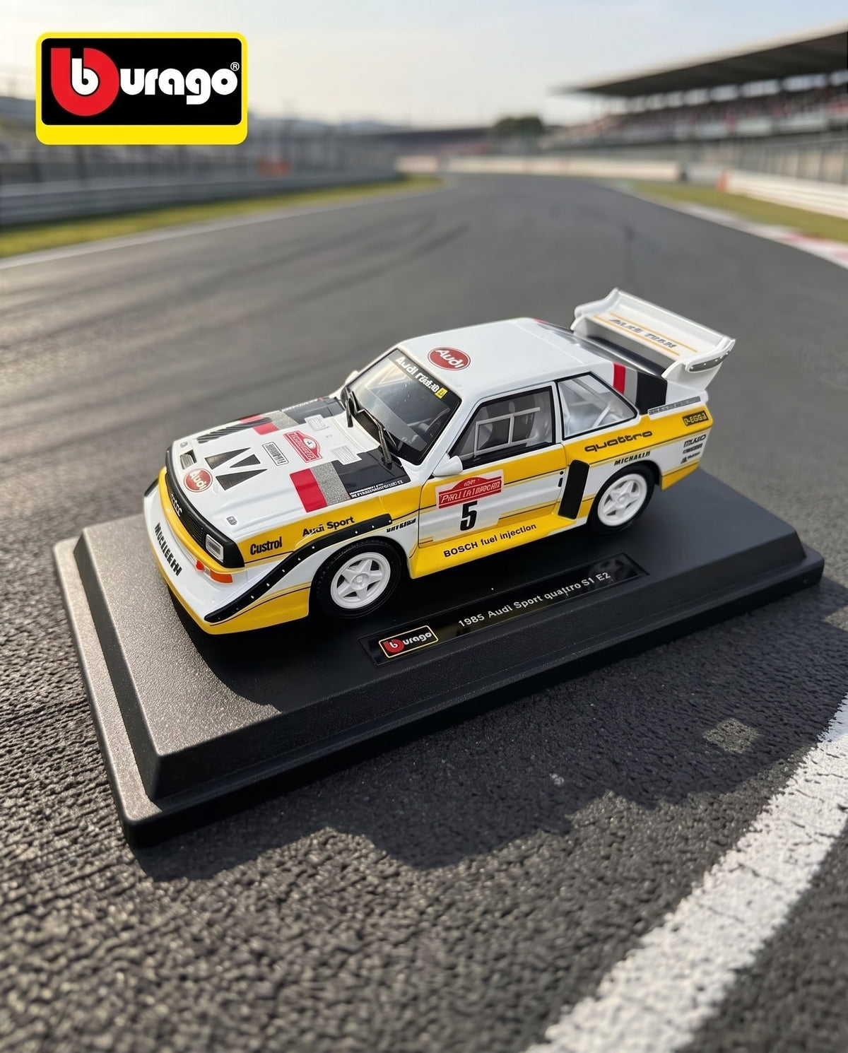 Official Bburago 1985 Audi Sport quattro S1 E2 diecast model car on stand with racetrack background, delivered nationwide across Lebanon.