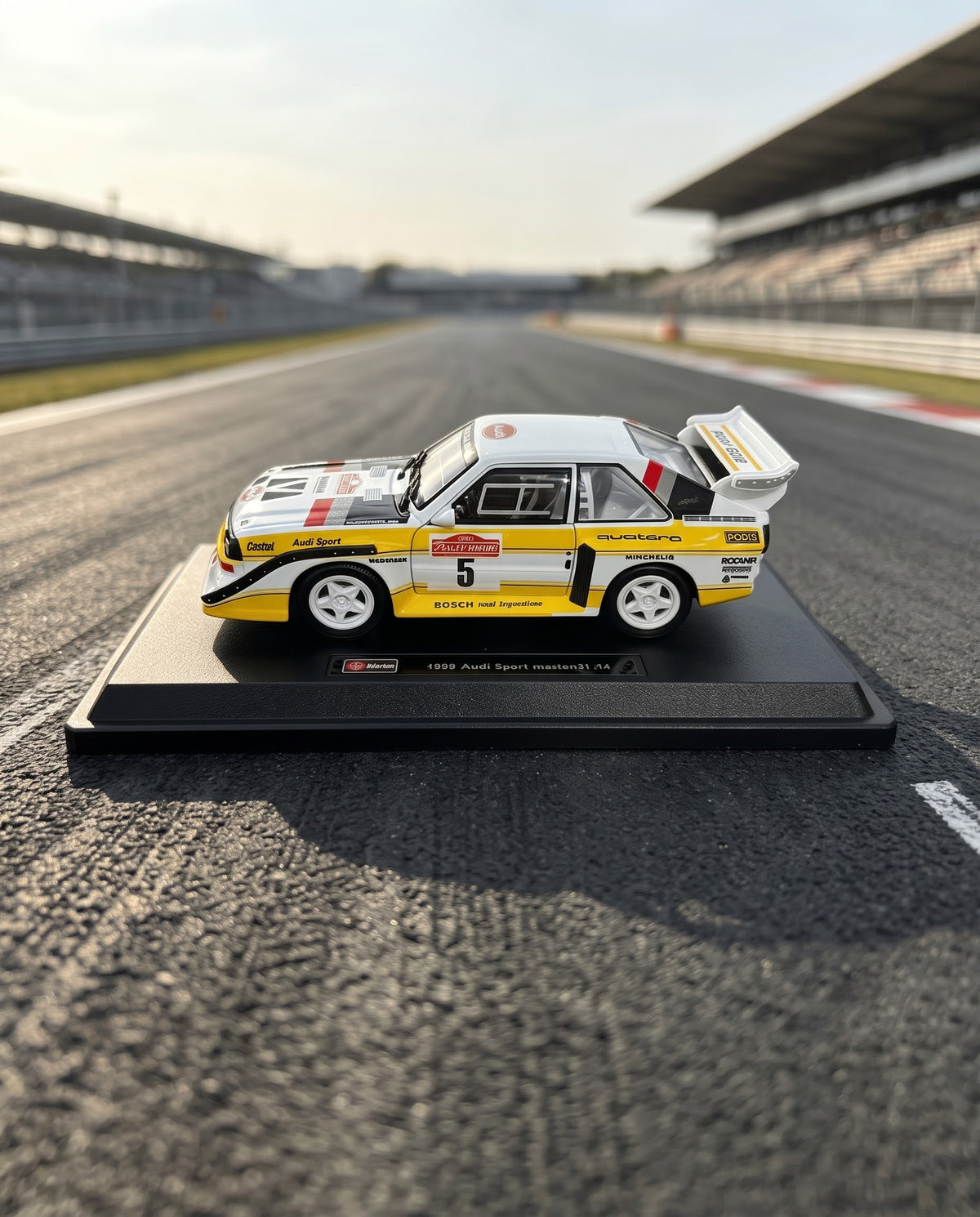 Full side view of 1985 Audi Sport quattro S1 E2 diecast collector car mounted on branded black display stand over track asphalt.