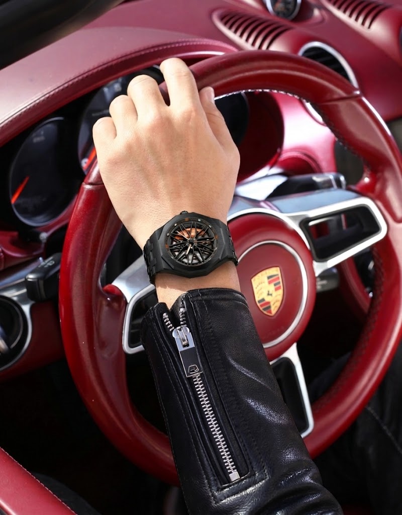 Black Poedagar spinning car wheel rim watch with orange caliper worn on wrist holding a sports car steering wheel.