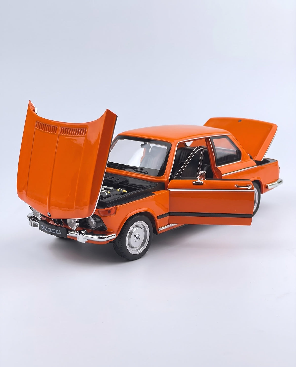 Front three-quarter view of the vintage orange BMW 2002 tii 1:18 scale model with open doors, trunk, and reverse-tilt hood, displaying front rally fog lights and chrome bumper.