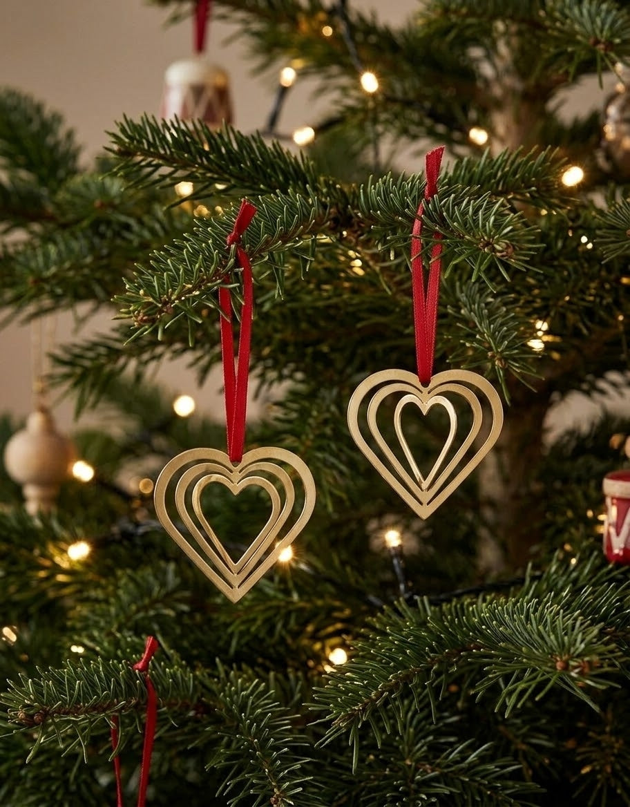 Set of layered gold metal heart ornaments hanging from spruce branches with festive holiday lights in background.