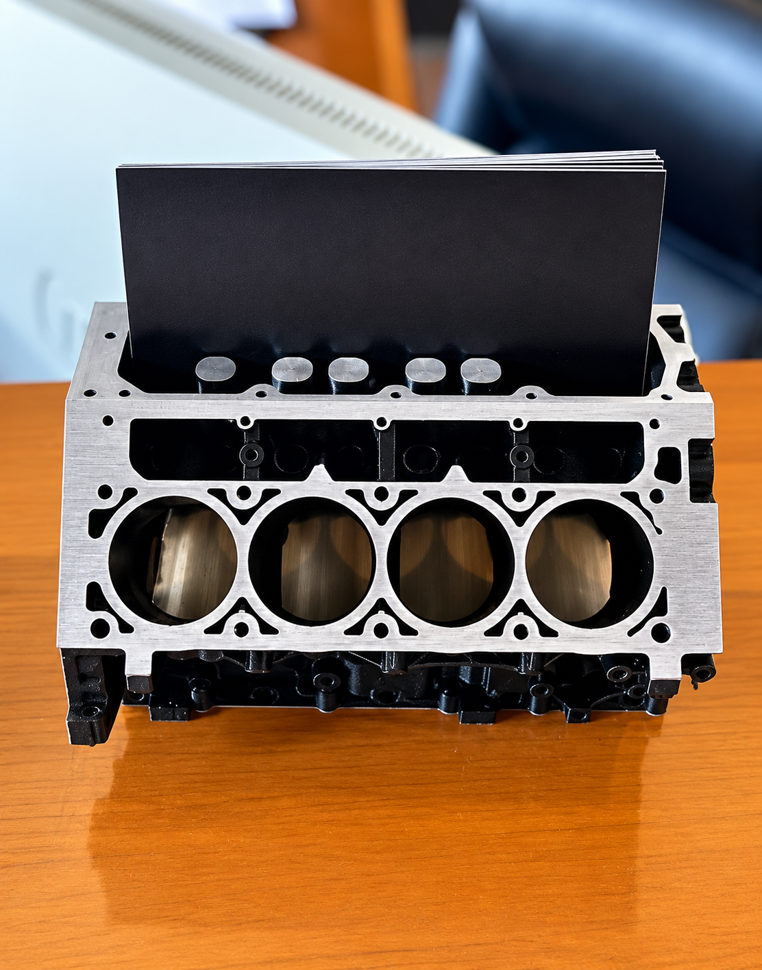 Front view of machined V8 engine block business card display stand on a glossy wooden desk surface.